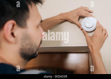Man installs white smoke detector on beige wall above door. Close-up view focuses on hands and device. Concept of home safety, fire prevention Stock Photo