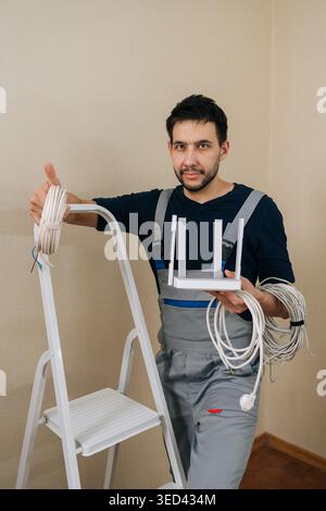 Service worker in overalls holding modern wireless router and network cables, giving thumbs up sign while standing next to ladder, ready to set up Stock Photo