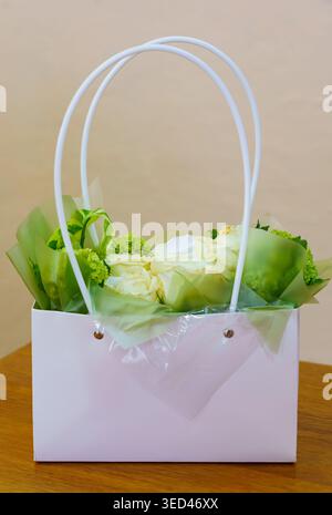 A white gift bag holds a bouquet of flowers with green wrapping. The setting is simple with a wooden table in a room. Stock Photo
