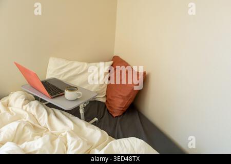 A laptop sits on a tray table next to a coffee cup in a corner of a bed covered with white blankets. Soft pillows lean against the wall in a simple se Stock Photo