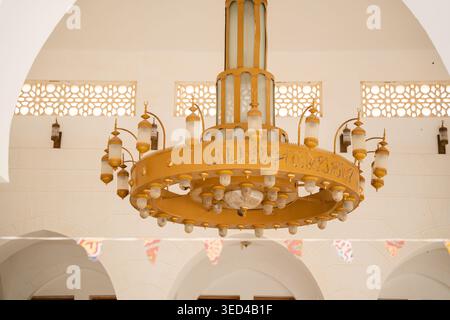 Large circular gold chandelier with multiple lamps and Arabic calligraphy hanging inside a mosque hall beneath white arches and patterned windows. Isl Stock Photo