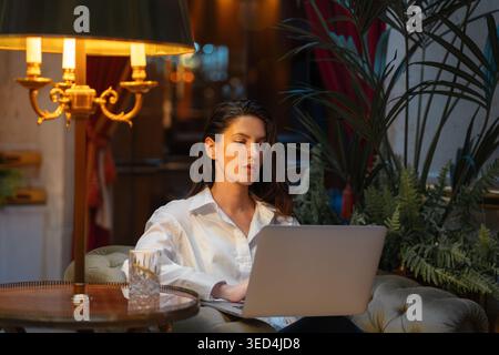 Focused businesswoman working on laptop in luxury hotel lobby with warm lighting and plants. Young woman in white shirt sitting on cushioned chair near glass of drink Stock Photo