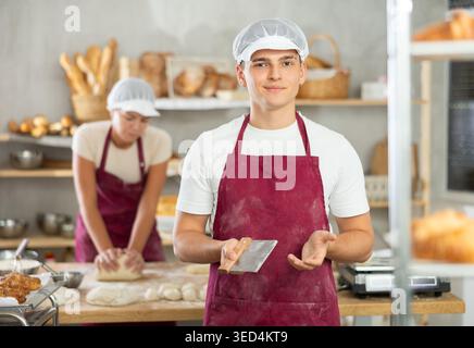 Young baker guy is standing near work table in kitchen, working with dough Stock Photo
