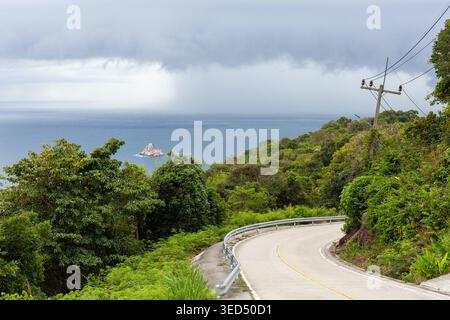Coastal road on Koh Tao, Thailand. Winding asphalt highway through lush green jungle. Dark monsoon storm clouds over the blue ocean. Stock Photo