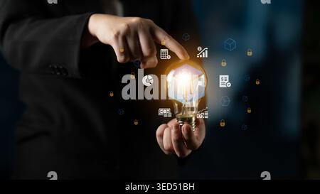 A business professional holds a glowing light bulb in one hand while pointing at digital icons, symbolizing innovation and creative ideas for future Stock Photo