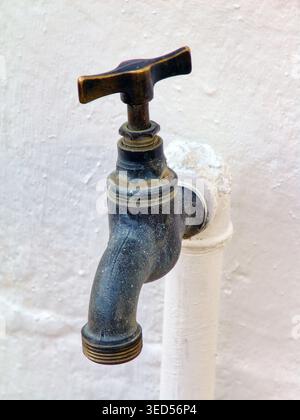 close-up shot shows a worn, dark metal outdoor faucet with a T-handle, mounted on a white-painted vertical pipe against a textured white wall. Stock Photo
