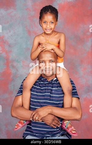 man in a striped shirt poses while holding his daughter on his shoulders against a mottled red and grey background. Stock Photo