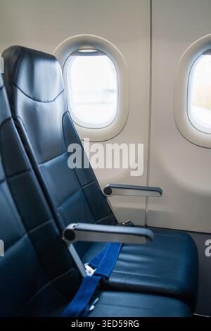 Two empty dark leather passenger seats with armrests and safety belts are located next to the bright windows inside a commercial aircraft cabin. Stock Photo