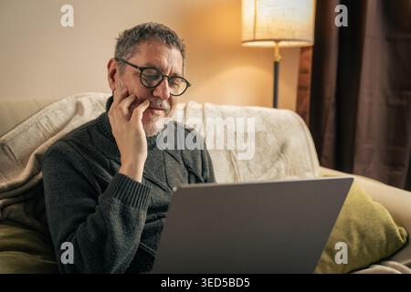 A middle-aged man with glasses and a beard sits on a comfortable couch at home, looking thoughtfully at his laptop screen while researching health and Stock Photo