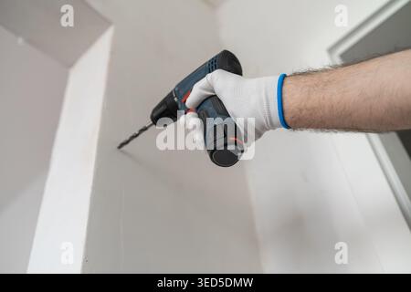 A technician using a cordless electric drill to install fixtures on a white wall. Clean and organized work environment with visible light fixture and tools in the background. Stock Photo