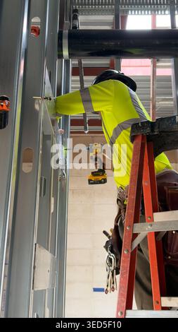 Construction worker installing metal framing using cordless drill on ladder indoors Stock Photo