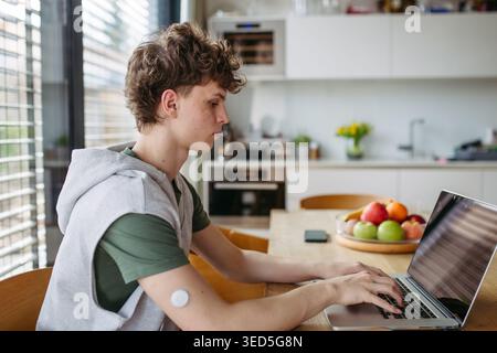 Teen boy with diabetes working on school project on laptop at kitchen table. Stock Photo