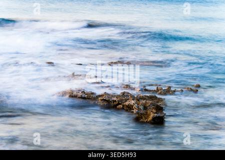 Long exposure seascape photograph capturing gentle ocean waves flowing around coastal rocks, creating a soft, dreamy motion blur in shades of blue and Stock Photo