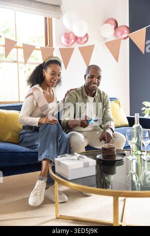 Couple sitting at home on blue sofa, man lighting cake candle with lighter, woman wearing sash Stock Photo