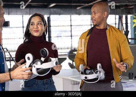 Diverse team standing open office maroon top and mustard jacket holding white headsets and tablet Stock Photo