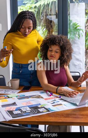 Two adult African American women collaborating at wooden table, arranging mockups and using laptop Stock Photo