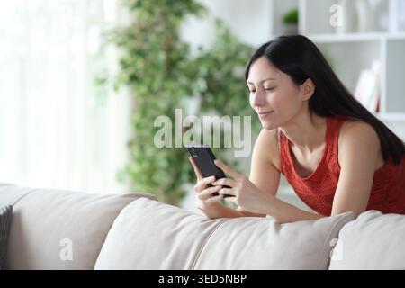 Asian woman using phone leaning on a couch at home Stock Photo