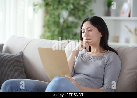 Thoughtful asian woman reading carefully a document sitting on a couch at home Stock Photo