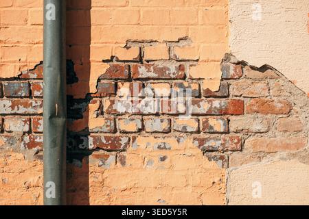 Weathered facade with flaking orange paint revealing warm brickwork beneath, textured masonry and gray metal pipe under strong daylight. Stock Photo