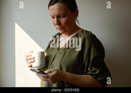 Side profile of middle-aged woman holding mug while texting on smartphone by window, warm morning sunlight and quiet home lifestyle moment, selective Stock Photo