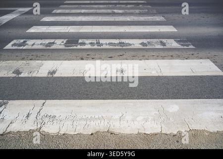 Texture of a weathered pedestrian zebra crossing on asphalt road. Thick white paint stripes heavily cracked and faded. Selective focus. Stock Photo