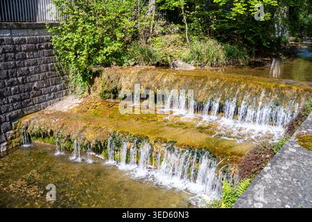 Small stepped waterfall over mossy rocks in a clear stream beside a stone retaining wall, with lush green trees and sunlight, tranquil nature scene. Stock Photo