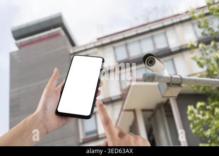Hand holding a blank smartphone near an outdoor CCTV camera and residential building, ideal for security apps, smart home monitoring, surveillance ser Stock Photo
