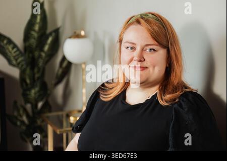 Woman with soft smile, houseplant and lamp in background indoors Stock Photo