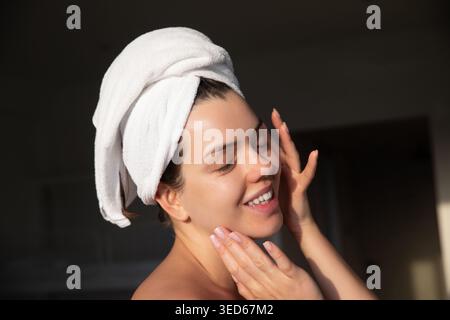 Pretty woman smiling gently. Hand touching cheek, smooth glowing skin, towel on head, relaxed posture. Beauty portrait, skincare concept. radiant heal Stock Photo
