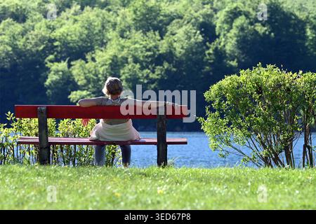 Senior woman relaxing on red park bench overlooking lake surrounded by green trees and shrubs, in Obertraun, Lake Hallstatt, Austria Stock Photo