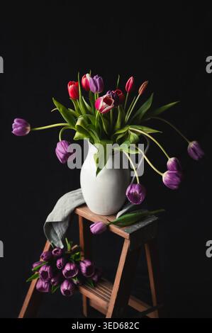 Jug of tulips on small wooden step ladder against black backdrop. Stock Photo