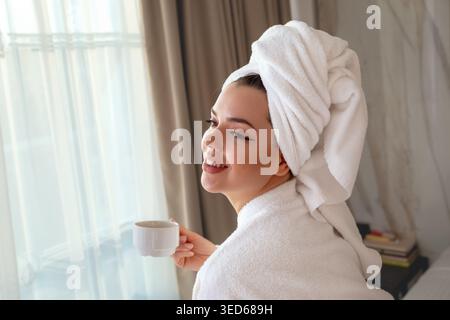 A woman smiles as she holds a cup of coffee while sitting by a window. Female is in a bathrobe and has a towel on her head. Soft light comes through t Stock Photo