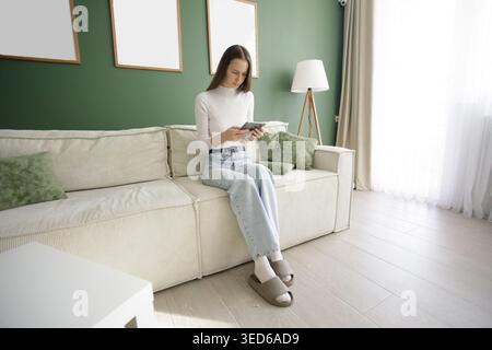 Lifestyle portrait of woman reading ebook gadget sitting on comfortable sofa in modern interior Stock Photo