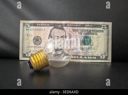 A conceptual image featuring a classic incandescent light bulb placed over a US five-dollar bill on a dark leather surface. This composition symbolize Stock Photo