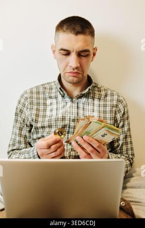 Man holds a bitcoin coin and euro banknotes while using a laptop at home. Concept of crypto investing, digital finance and comparing traditional money Stock Photo