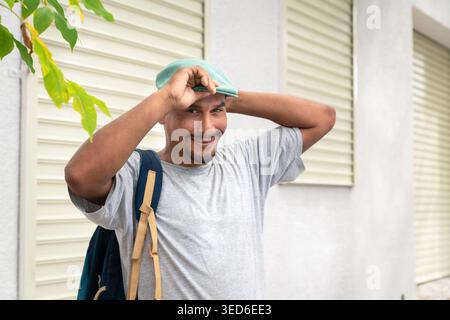 Portrait of a young Latin Hispanic man with backpack adjusting his cap while standing on a residential city street. Stock Photo
