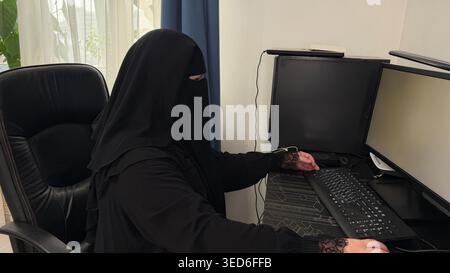 Woman wearing hijab using desktop computer with empty white monitor. Concept of web design, UI mockup and digital services. Stock Photo