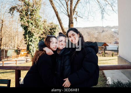 Three young Caucasian sisters standing together and hugging outdoors in park setting smiling at camera. Family relationship and lifestyle. Stock Photo