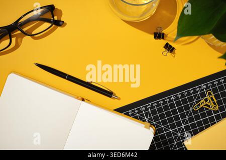 Open notebook with black pen on bright yellow desk. Glasses, binder clips, paper clips, cutting mat. Copyspace Stock Photo