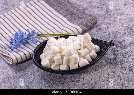Fluffy white mini marshmallows fill black bowl. Striped grey, white napkin, delicate blue spring flowers rest beside Stock Photo