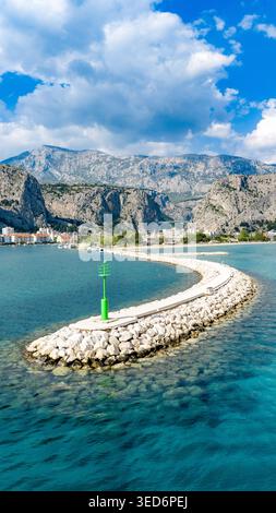Aerial view of the curved stone breakwater and green beacon in the turquoise Adriatic Sea with the coastal town and rugged mountains in the background Stock Photo