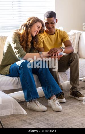 Diverse couple sitting on light sofa in bright living room, holding hands and wearing smartwatches Stock Photo