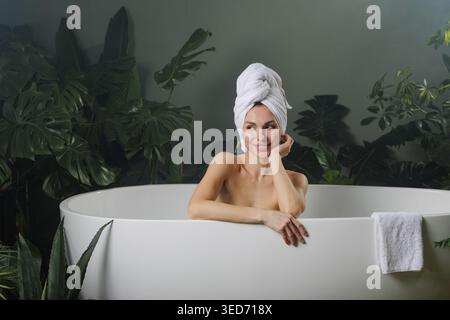 A serene woman with a towel on her head enjoys a moment of relaxation in a modern bathtub, surrounded by tropical plants, perfect for wellness and bea Stock Photo
