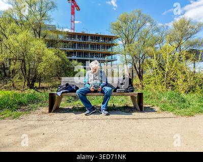 Elderly gray-haired man sitting on a park bench, focused on operating a drone with a handheld drone controller. Sunny spring. Trees and building workp Stock Photo