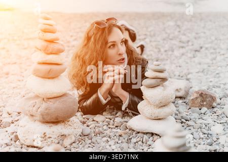 Woman beach stones lying on a pebble shore with stacked rock towers while gazing off into the distance during sunset Stock Photo