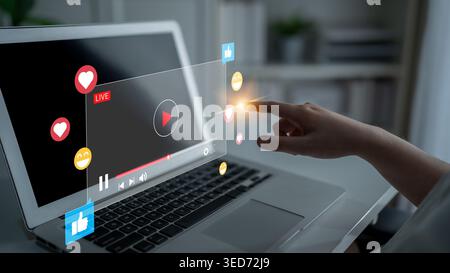 A focused hand interacts with a laptop displaying a live streaming interface, surrounded by social media reaction icons, representing modern digital Stock Photo