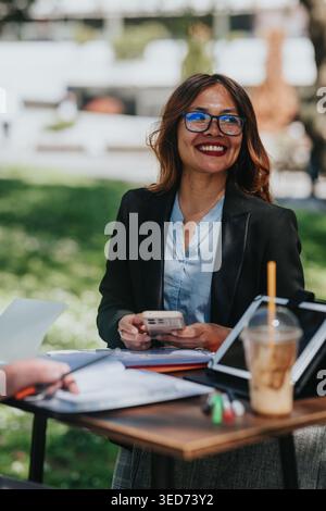 Smiling businesswoman using smartphone during outdoor meeting with tablet and coffee Stock Photo