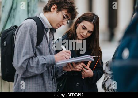 Two students studying together outdoors taking notes and discussing on a city campus Stock Photo