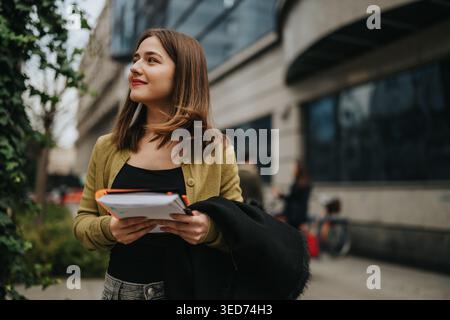 Young student holding notebooks outdoors on a modern urban campus, looking thoughtful and confident Stock Photo