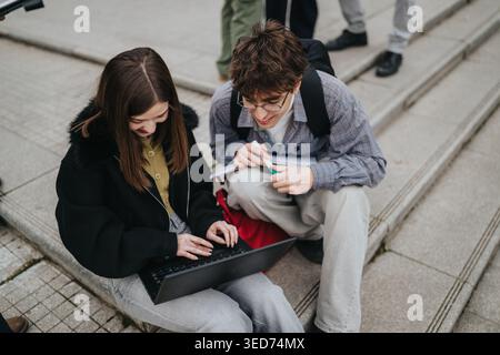 Two students learning together on outdoor steps with laptop and notebooks, collaborating and smiling Stock Photo